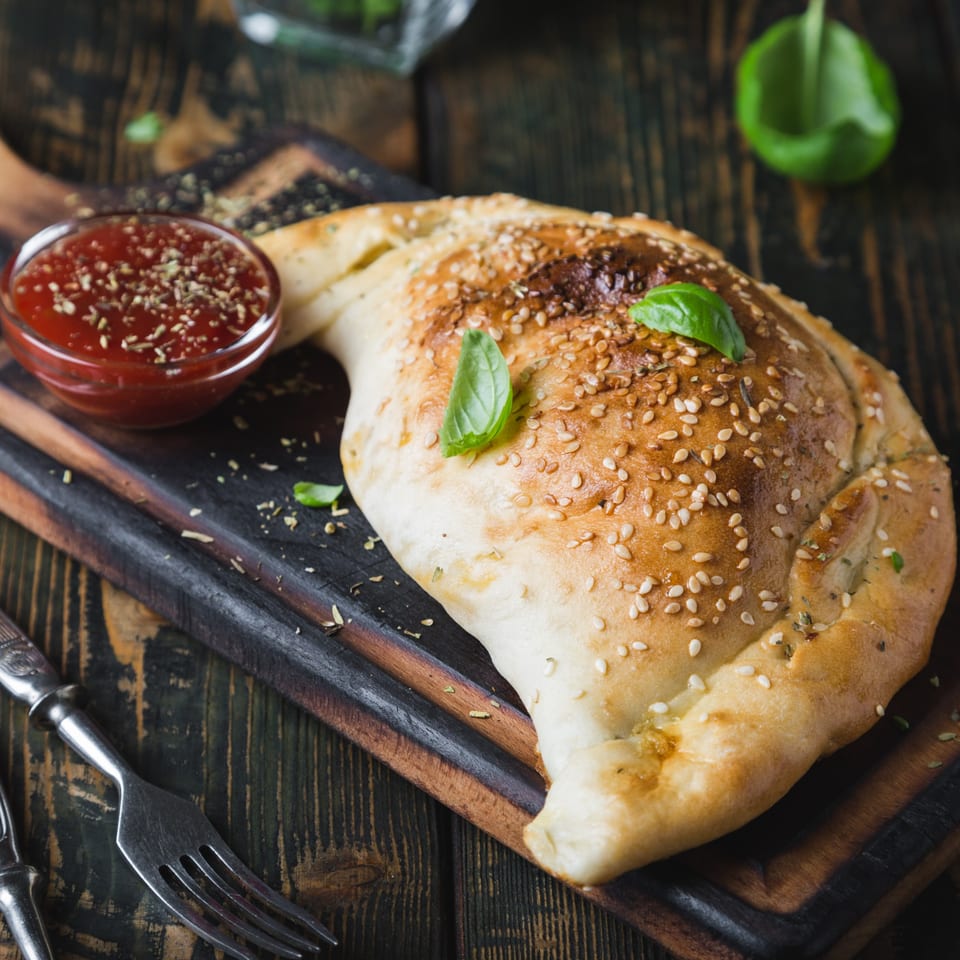 A golden-brown Speedy's Pizza calzone, topped with sesame seeds and basil, served with marinara sauce.
